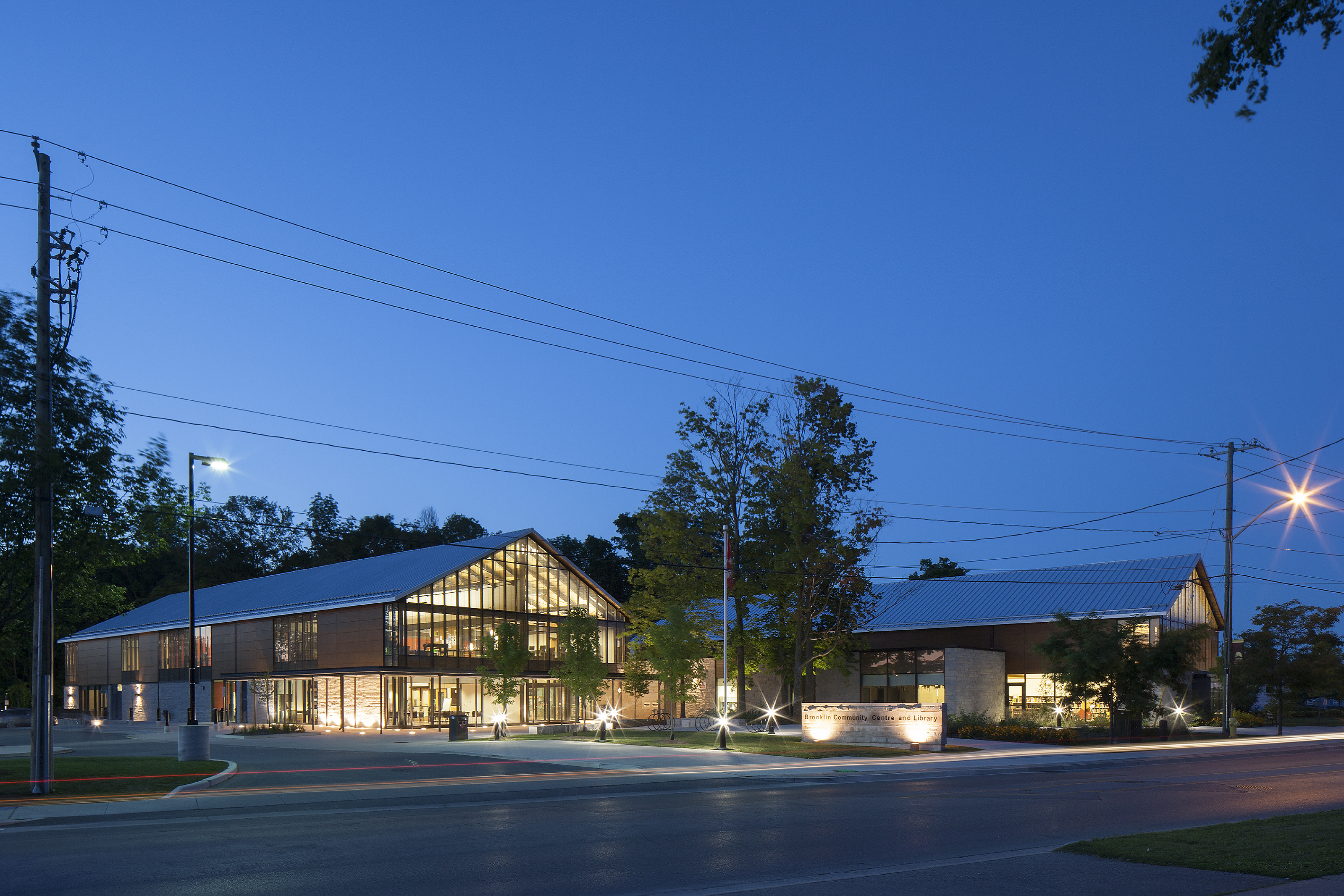 Two large, low, interconnected buildings with metal gable roofs and glass and stone walls. The buildings glow with warm light against an evening sky highlighting the sillouhette of trees around them.